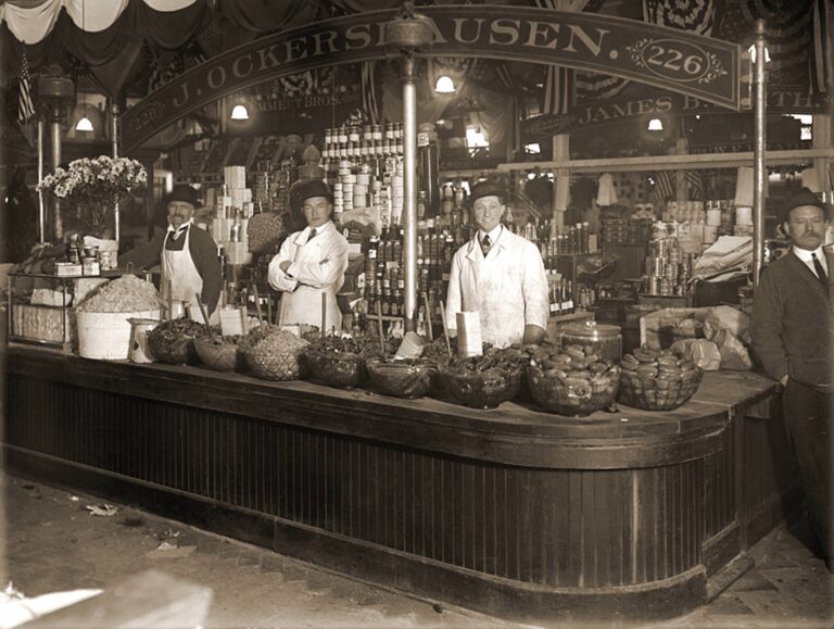 Vendors stand behind a counter displaying an assortment of pickles and other food goods in a vintage photograph