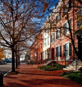 A tree-lined street features a neat row of brick homes under a bright blue autumn sky